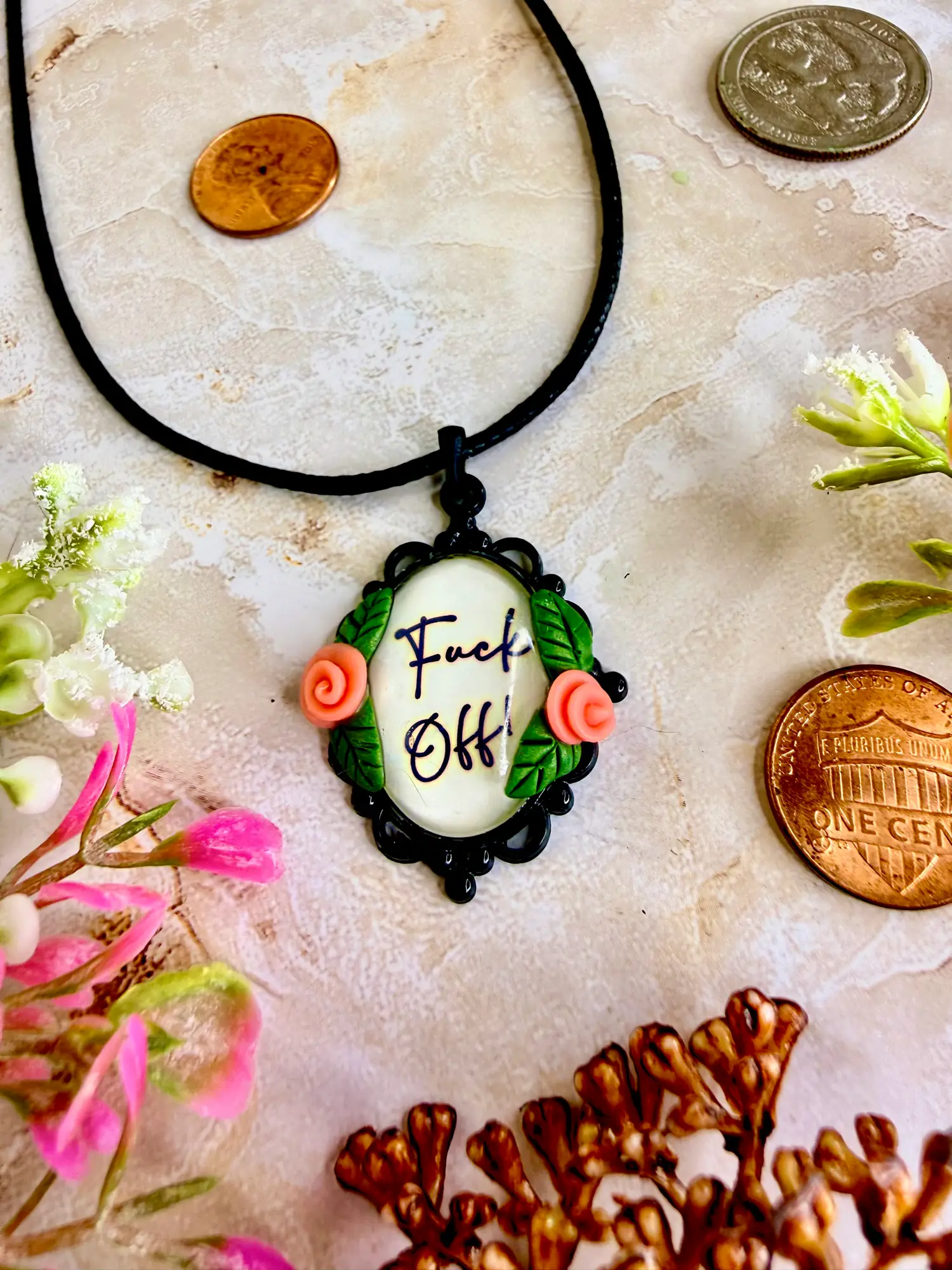 Fuck off written in fancy letters and embedded into a black cameo pendant with spiral pink flowers and placed on a faux leather black cord