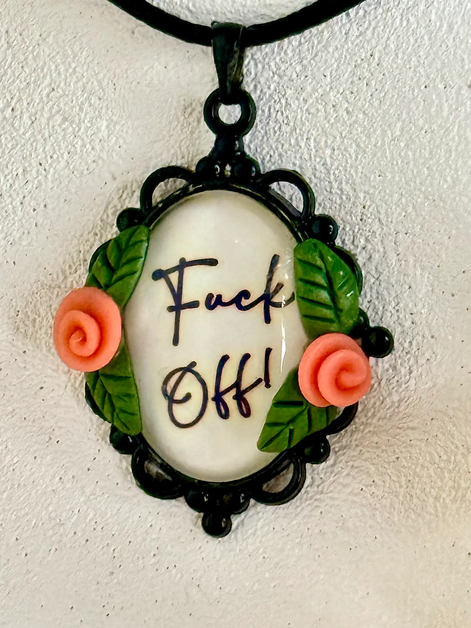 Fuck off written in fancy letters and embedded into a black cameo pendant with spiral pink flowers and placed on a faux leather black cord